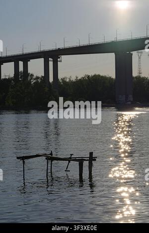 Moderno ponte autostradale in cemento che si estende su acque di fiume calme con un sentiero di sole scintillante da un sole luminoso che riflette la luce sulla superficie, forma Foto Stock