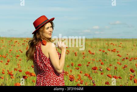 Bella donna dai capelli rossi che guarda indietro con un sorriso gentile mentre indossa un cappello rosso e un vestito rosso in un campo vibrante di papaveri e orzo sotto un cielo blu. Foto Stock