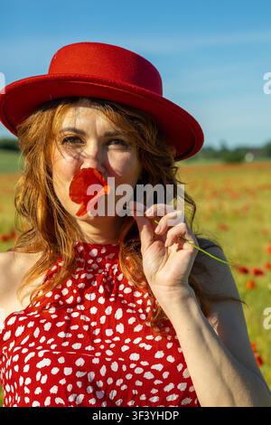 Ritratto ravvicinato di una donna allegra dai capelli rossi che indossa un cappello rosso e un vestito a pois rossi mentre tiene un fiore di papavero rosso acceso vicino al viso. Foto Stock
