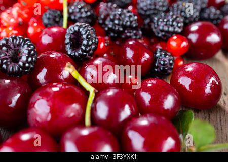 Vista macro di ciliegie scure mature mescolate con lamponi neri e ribes rosse su una tavola di legno rustica, che mostra una pelle lucida e bagnata, un contrasto di colori vivaci, una profondità di campo ridotta e una freschezza naturale Foto Stock