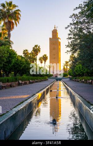 Il minareto della Koutoubia si erge sopra gli alberi a Marrakesh. Il sole tramonta, proiettando una luce calda sull'edificio. Un canale d'acqua calmo riflette la torre e i giardini circostanti. Foto Stock