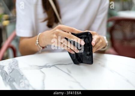 Donna che tiene un portafoglio nero a un tavolo da caffè all'aperto. Primo piano di una donna che tiene un portafoglio nero strutturato sopra un tavolo di marmo mentre è seduta a un tavolo all'aperto Foto Stock