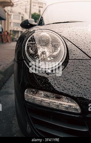 Dettaglio del faro anteriore e degli indicatori di direzione di una Porsche 911 (997) nera ricoperta di gocce di pioggia, parcheggiata su una strada cittadina Foto Stock