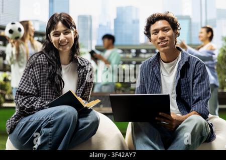 Ritratto di studenti diversificati che sorridono alla telecamera mentre studiano con laptop e libro aperto, seduti su un pouf nel moderno campus. Studio accademico internazionale. Foto Stock