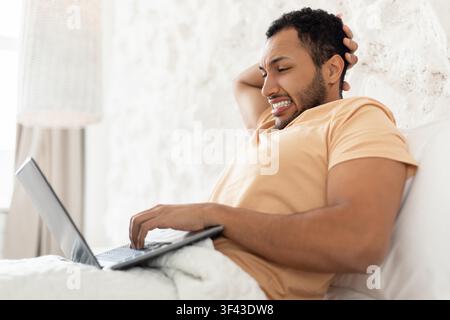 Uomo nero felice che lavora su un laptop mentre si rilassa a letto, concetto di ufficio domestico Foto Stock