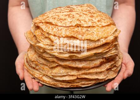 Prima piano di una pila enorme di pancake tradizionali fatti in casa presentati nelle mani su uno sfondo nero Foto Stock