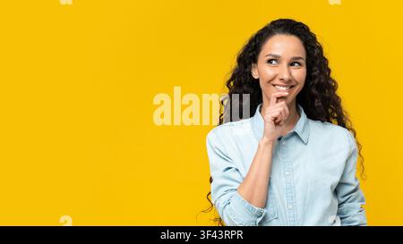 Giovane donna pensierosa con capelli ricci che sorride e guarda verso l'alto, sfondo giallo Foto Stock