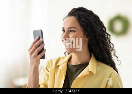Giovane donna latina sorridente che tiene uno smartphone e partecipa a una videochiamata a casa Foto Stock