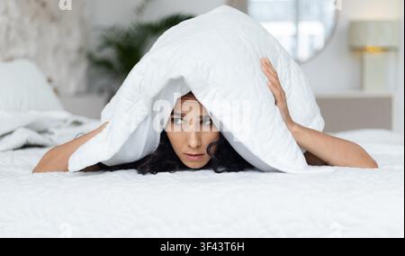 Giovane donna preoccupata che si nasconde sotto la coperta nella camera da letto, stato emotivo di ansia e paura Foto Stock