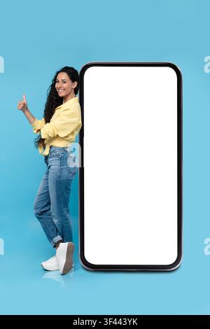 Giovane donna ispanica sorridente che dà il pollice alzato accanto a uno smartphone vuoto, concetto di mockup Foto Stock