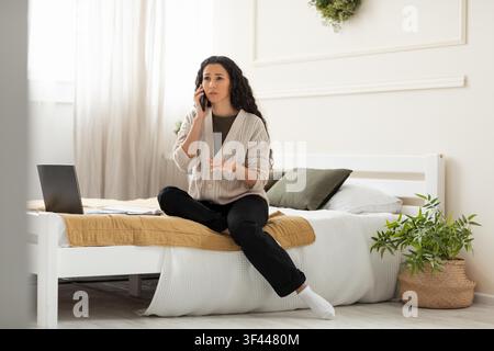 Donna ispanica preoccupata che parla al telefono seduta sul letto a casa, situazione stressante Foto Stock