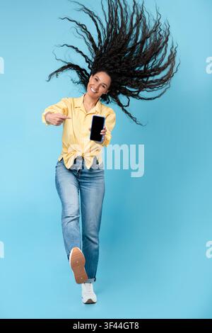 Giovane donna sorridente con capelli ricci che mostra uno smartphone, sfondo blu, concetto di tecnologia Foto Stock