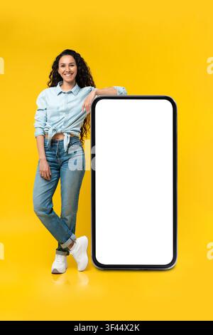 Giovane donna ispanica sorridente appoggiata a uno smartphone con schermo vuoto, sfondo giallo Foto Stock