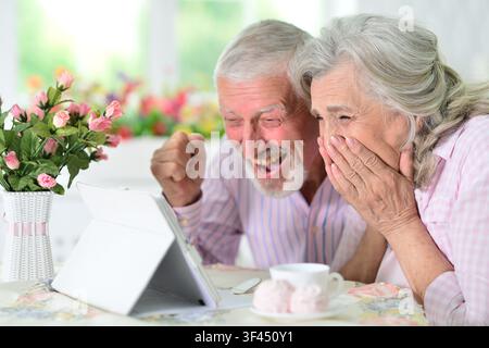 anziano gioioso che ride insieme davanti a un tablet in un ambiente domestico con fiori rosa e bianchi in un vaso e tazza in ceramica con piattino su sfondo tessile. moderno Foto Stock