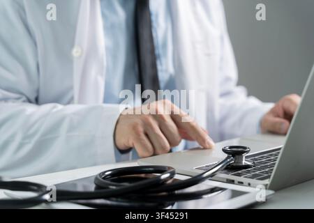 Primo piano di un medico che lavora su un laptop accanto a uno stetoscopio e un tablet, rappresentante la telemedicina, la consultazione online, l'assistenza sanitaria digitale, la medicina Foto Stock