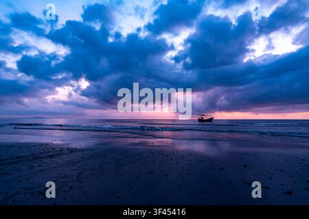Una scena serena dell'alba tropicale con una barca da pesca tradizionale silouettata contro un cielo drammatico con nuvole blu, viola e rosa vibranti, re Foto Stock