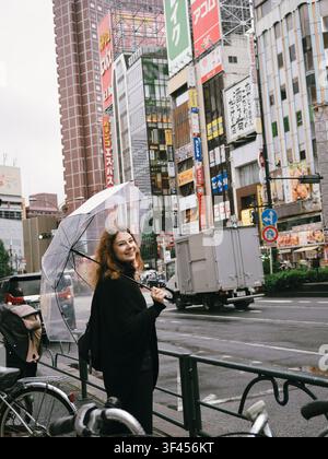 Giovane donna che tiene un ombrello trasparente su una strada con cartelloni pubblicitari in una giornata di pioggia a Shinjuku, Tokyo, Giappone Foto Stock