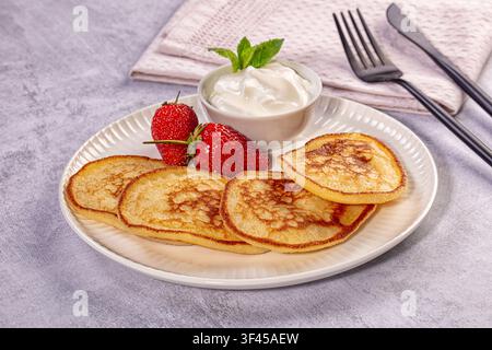 Pancake dorati disposti su piatto bianco con fragole rosse fresche e panna montata, guarniti con menta Foto Stock