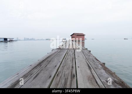 Un molo di legno stagionato si estende sopra lo stretto di Penang verso una piccola casa su palafitte di legno rosso costruita sull'acqua, con barche da pesca e un profilo urbano sfocato visibile sullo sfondo. Foto Stock