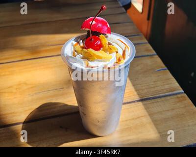 Milkshake decadente in una tazza trasparente sormontata da panna montata, caramello e una ciliegia su un tavolo di legno rustico alla luce naturale. Foto Stock