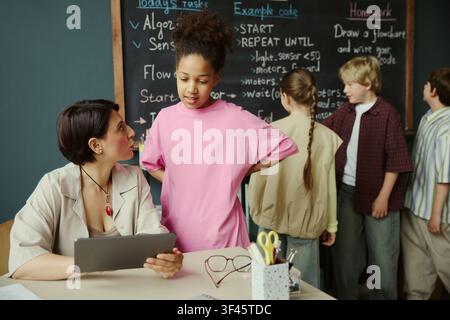 Studentessa nera discute un compito di programmazione con un'insegnante mentre i compagni di classe lavorano alla lavagna, mostrando apprendimento STEM e mentoring. Utile per l'educazione, lezioni di programmazione, campagne sulle competenze digitali Foto Stock