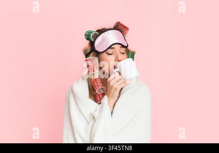Donna assonnata in arricciacapelli e maschera per il sonno avvolto in coperta e bevande caffè da tazza Foto Stock