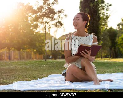 Donna asiatica si gode la lettura di un libro in un parco in una giornata estiva. Foto Stock