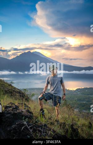 Giovane fotografo su una cresta erbosa che si affaccia sul Monte Batur e sul Lago Batur all'alba a Kintamani, Bali, Indonesia. Foto Stock