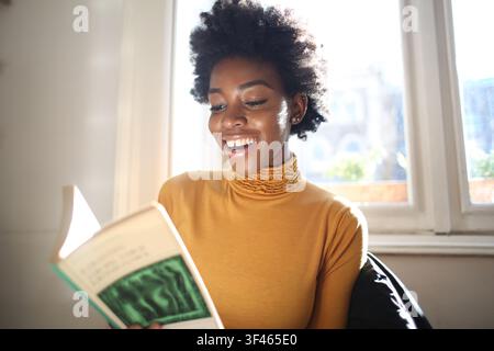 Giovane donna gioiosa che si gode un libro vicino alla finestra Foto Stock