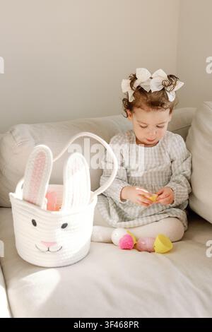 Bambina apre uova di Pasqua colorate accanto a un cestino con coniglietto a casa Foto Stock