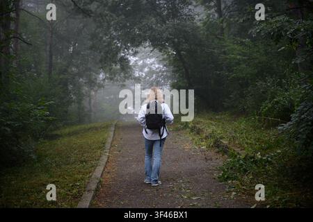 Una donna con capelli biondi si allontana dalla fotocamera su un sentiero asfaltato attraverso una foresta densa e nebbiosa. Indossa una camicia bianca a bottoni, b Foto Stock