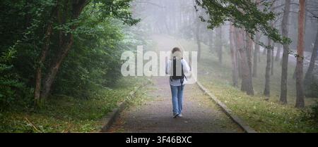 Una fotografia orizzontale di una donna che si allontana dall'osservatore su un sentiero lastricato attraverso una foresta nebbiosa. È vestita in modo casual con blue jeans e un l Foto Stock