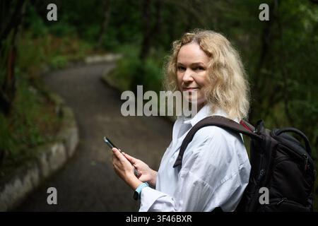 Una donna caucasica di mezza età con capelli biondi ricci si trova su un sentiero lastricato in un'area boschiva, guardando indietro sopra la spalla verso la fotocamera. Indossa Foto Stock