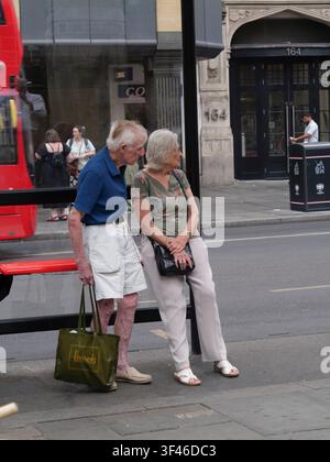 14 luglio 2026. Una coppia anziana aspetta pazientemente a una fermata dell'autobus su Bishopsgate nella City di Londra, UK, con l'uomo vestito con pantaloncini estivi e che tiene una borsa della spesa distintiva verde del grande magazzino Harrods durante il recente periodo di caldo Foto Stock