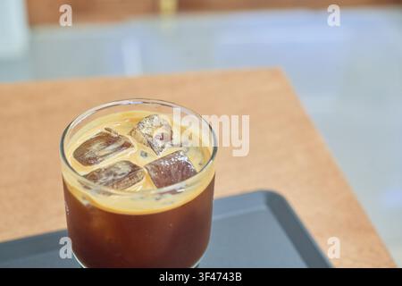 Bicchiere trasparente di caffè freddo con cubetti ghiacciati e crema dorata su un tavolo di legno in un caffè moderno che evidenzia stili di vita urbani quotidiani rilassanti. Foto Stock
