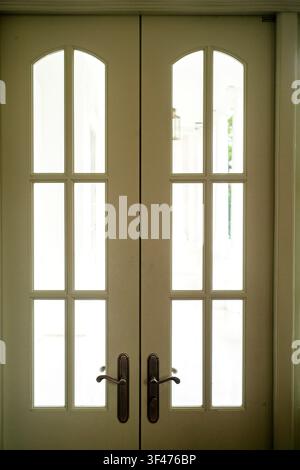 Eleganti porte doppie francesi bianche con vetri a vetri arcuati e maniglie classiche in bronzo che conducono in uno spazio interno luminoso. Foto Stock