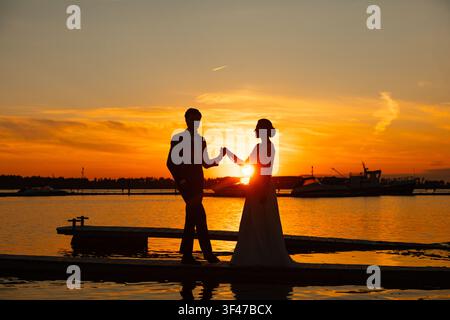 Sposa e sposo che si tengono per mano al tramonto vicino al lago, silhouette del matrimonio Foto Stock