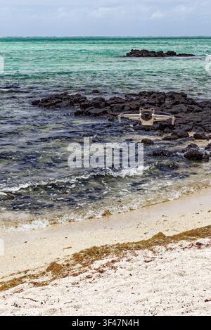 Scatto verticale all'altezza degli occhi che cattura un moderno drone quadrocopter in volo stabile a mezz'aria sopra una spiaggia tropicale accidentata. Located at Ile aux Cerf Foto Stock