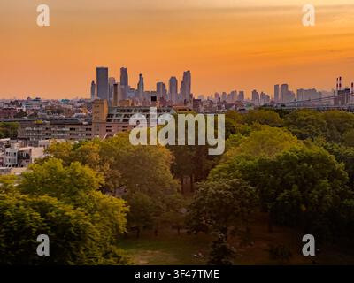 Un tramonto dorato illumina lo skyline di Manhattan visto da Brooklyn con una lussureggiante area verde in primo piano e un ponte che attraversa l'East River. Foto Stock