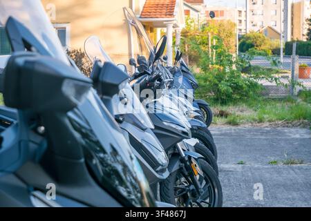 Filiera di scooter grigi parcheggiati ordinatamente lungo un marciapiede in una zona residenziale con piante verdi ed edifici sullo sfondo. Foto Stock