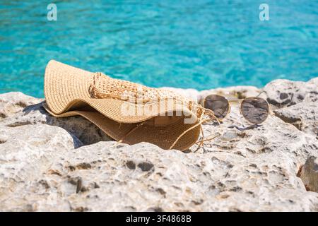 Un cappello di paglia e occhiali da sole appoggiati su una superficie rocciosa vicino all'acqua limpida e blu. Ideali per temi estivi e da spiaggia. Foto Stock
