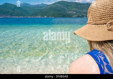 Una donna con lunghi capelli biondi è seduta accanto al mare blu chiaro, indossando un cappello di paglia. Lo sfondo mostra montagne e un cielo soleggiato. Foto Stock