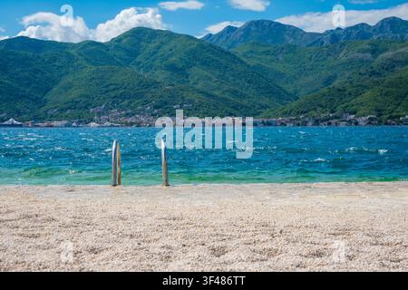 Spiaggia di ciottoli con ringhiere metalliche per l'accesso all'acqua, acqua blu limpida e onde dolci, sfondo di montagne verdi lussureggianti, scena costiera serena. Foto Stock