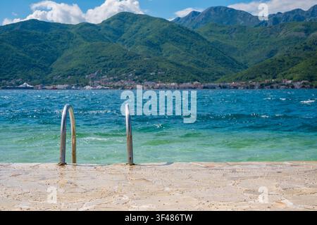 Spiaggia di ciottoli con ringhiere metalliche per l'accesso all'acqua, acqua blu limpida e sfondo montano, scena serena di vacanza costiera. Foto Stock