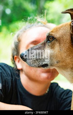 Primo piano del profilo laterale di un cane marrone che guarda avanti, con il proprietario leggermente sfocato sullo sfondo, all'aperto con luce naturale e bokeh verde e ca Foto Stock
