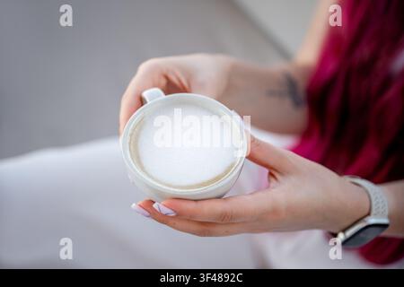 Primo piano delle mani di una donna che tiene una tazza bianca di cappuccino con schiuma di latte cremosa. Momento accogliente in un caffè con luce soffusa, abbigliamento casual e vibrazioni rilassanti Foto Stock