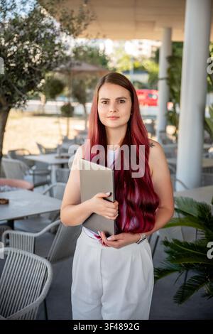 Giovane donna professionista sicura di sé con lunghi capelli rosa tiene un laptop su una terrazza all'aperto di un caffè, suggerendo lavoro a distanza, freelance, incontro di lavoro Foto Stock