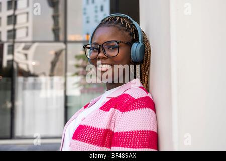 Donna nera sorridente con trecce e occhiali, che indossa cuffie sopra l'orecchio, appoggiata a un muro in città, godendosi la musica durante un viaggio urbano Foto Stock