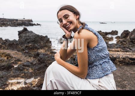 Una donna sorridente siede pacificamente sulla costa vulcanica di Lanzarote. Indossa occhiali e un top floreale blu, con l'oceano e formazioni rocciose in th Foto Stock