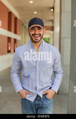 Giovane uomo allegro con cappellino da baseball e camicia azzurra abbottonata sorride all'aperto in un corridoio moderno, mani in tasca, luce naturale e s Foto Stock
